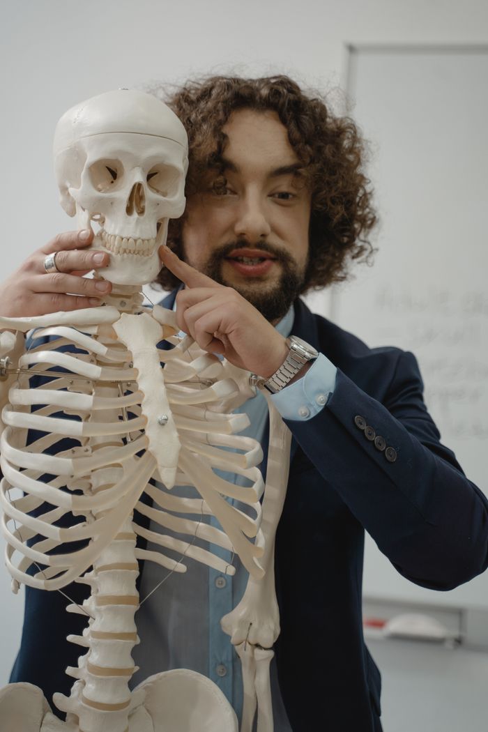 Male teacher in a suit explains a human skeleton model in a classroom setting, pointing at the skull.