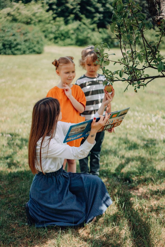 A teacher reads to children under a tree, promoting outdoor education.