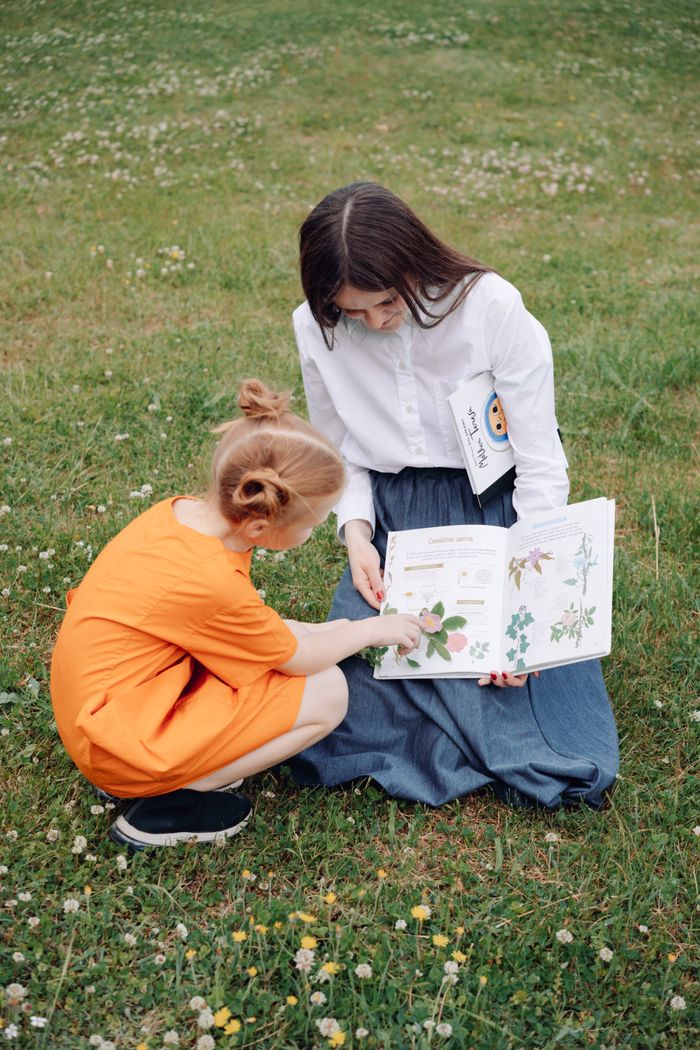 A child and teacher explore a nature book together while sitting on grass in a field.